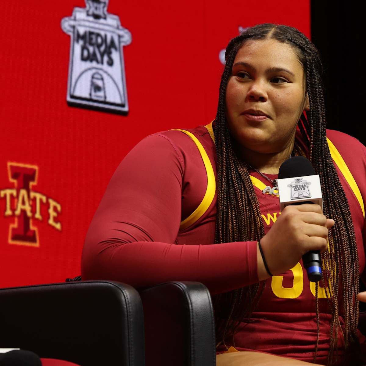 Female college athlete in a red and gold jersey speaks into a microphone at a media day, with a red backdrop and logos behind her.
