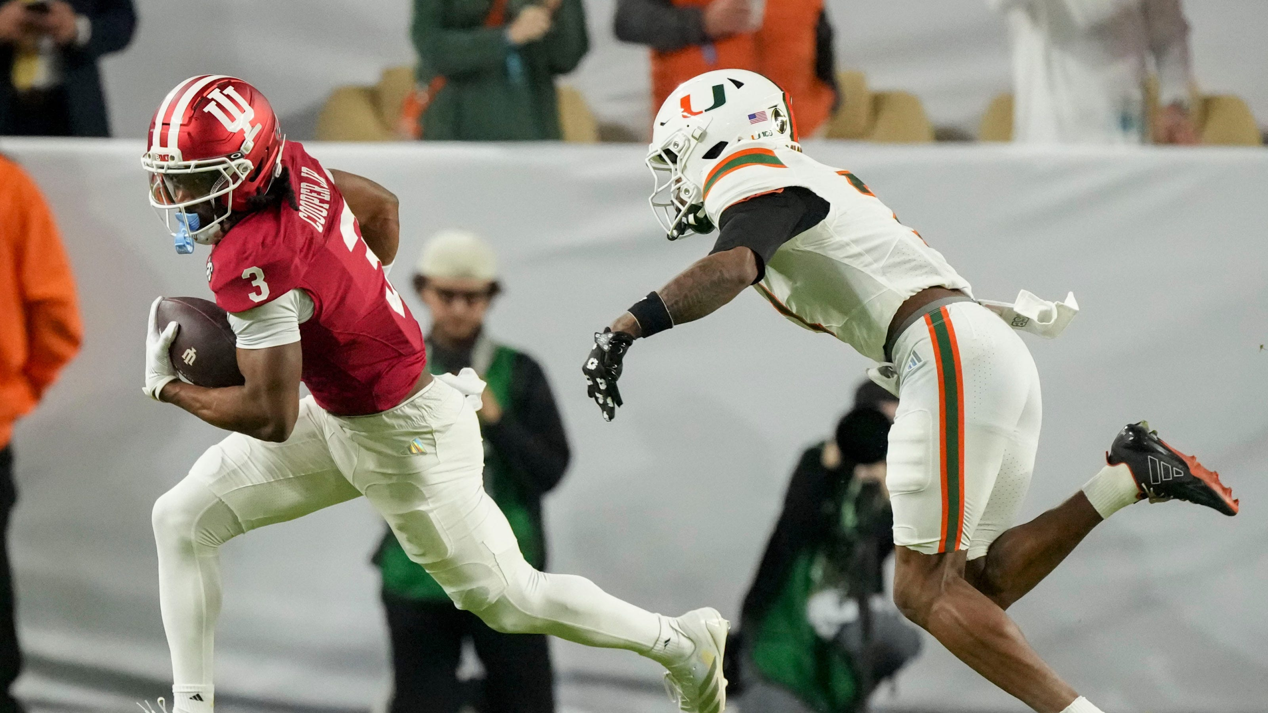 Alabama running back in a red jersey carries the ball while sprinting past a Miami defender in white during a college football game.
