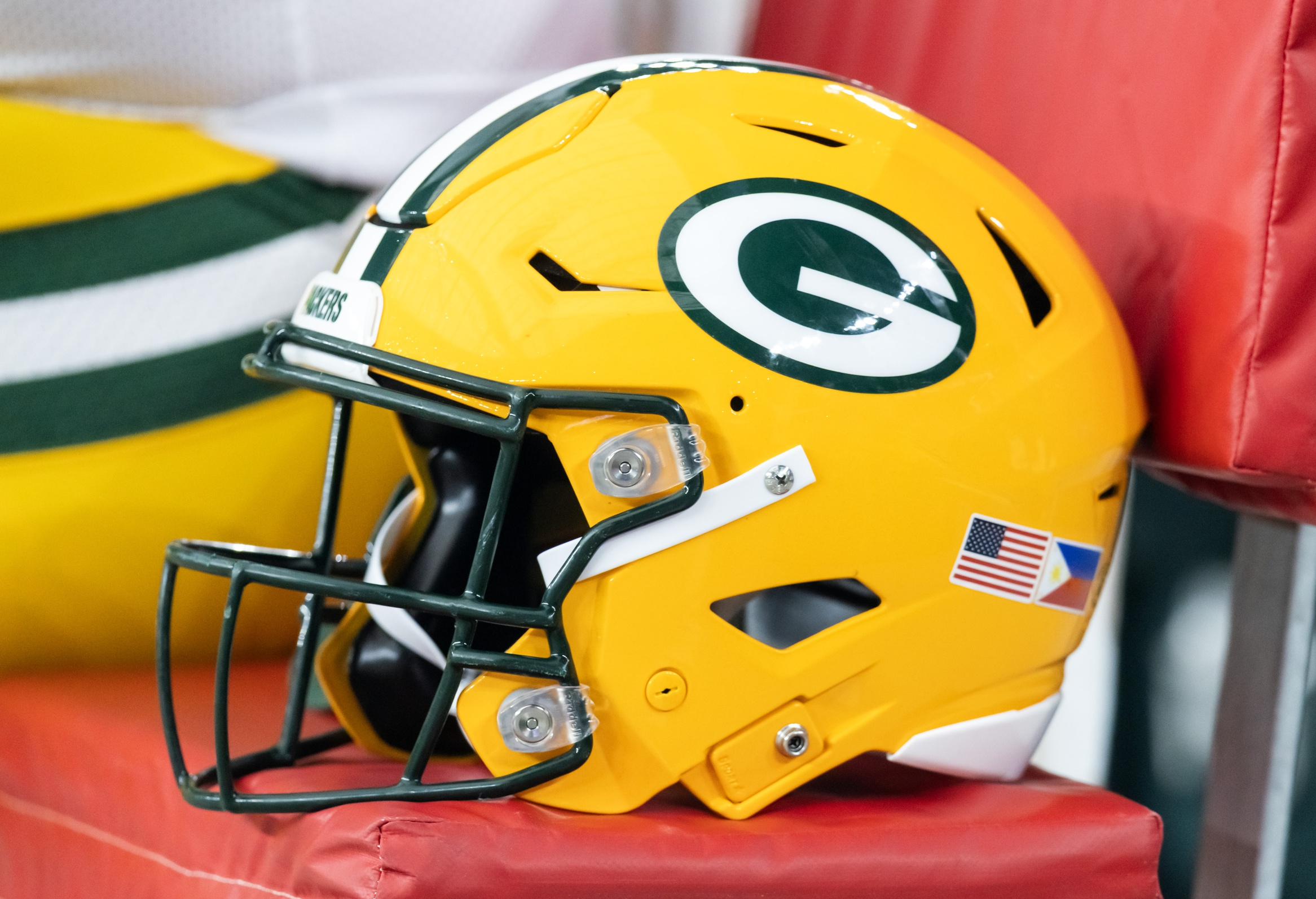 Green Bay Packers football helmet in gold with a dark green faceguard resting on a red padded surface, showing the Packers 'G' logo on the side and small sticker decals.